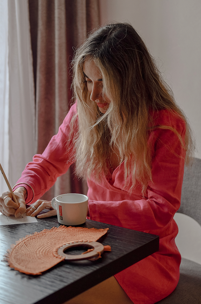 A blonde girl sitting in a desk writing in a note pad with a circular orange palm fan in front of her.
