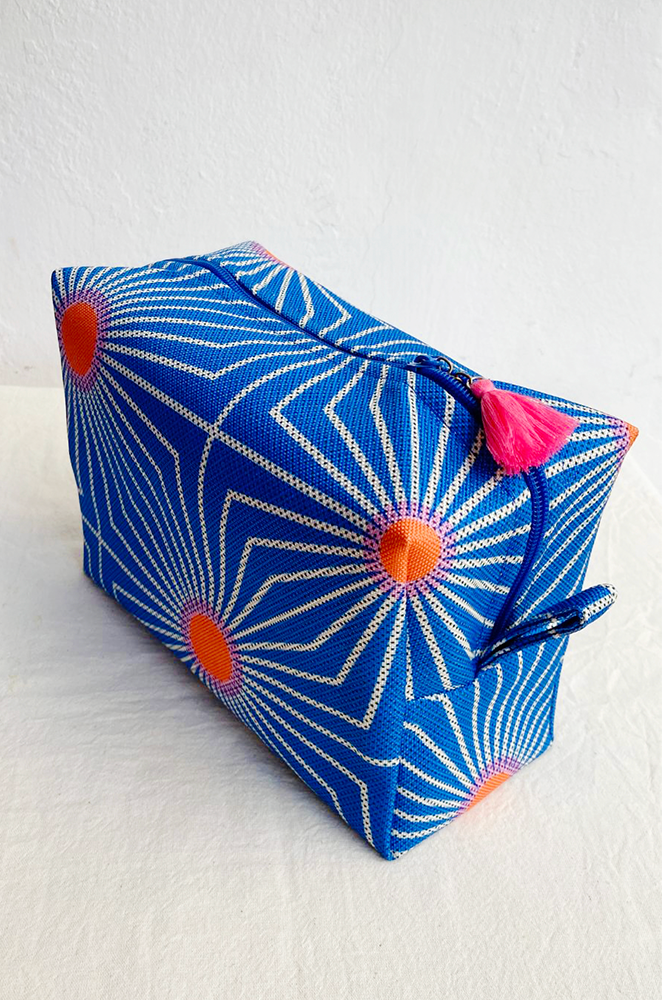 side view of a travel toiletry bag with a little pink charm in the zipper. The print is blue with a geometric sun pattern.