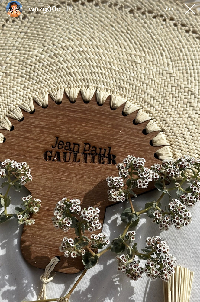 Branded wooden fan with Jean Paul Gaultier logo. Surrounded by little white flowers. 
